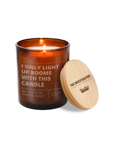 An amber glass candle jar with a lit flame and a light wood lid leaning against it on a white background. The jar has white text reading 'I ONLY LIGHT UP ROOMS WITH THIS CANDLE' and the lid is engraved with 'THE INVESTIGATION' and the WinShop brand.