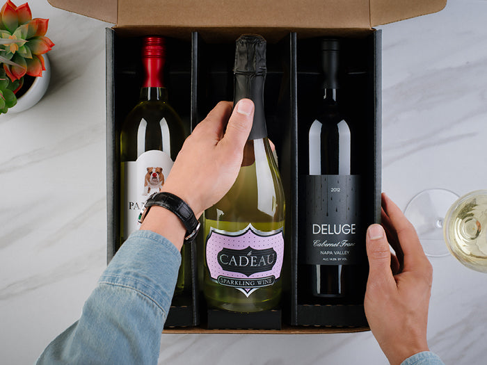 Person selecting a bottle of wine from a box with other bottles on a table.