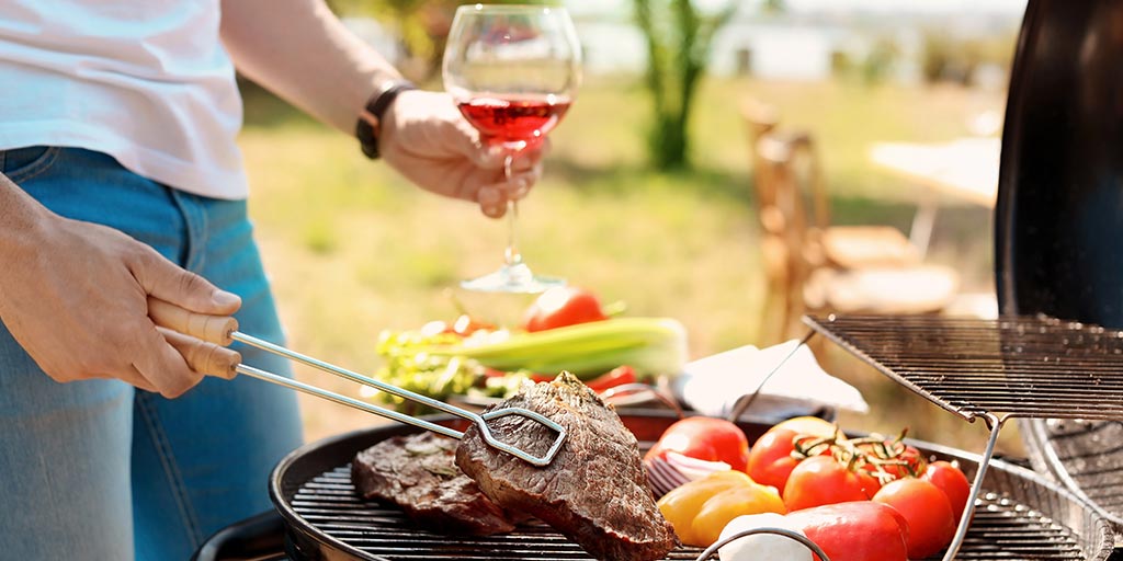 Man grilling steak and vegetables outdoors with a glass of wine