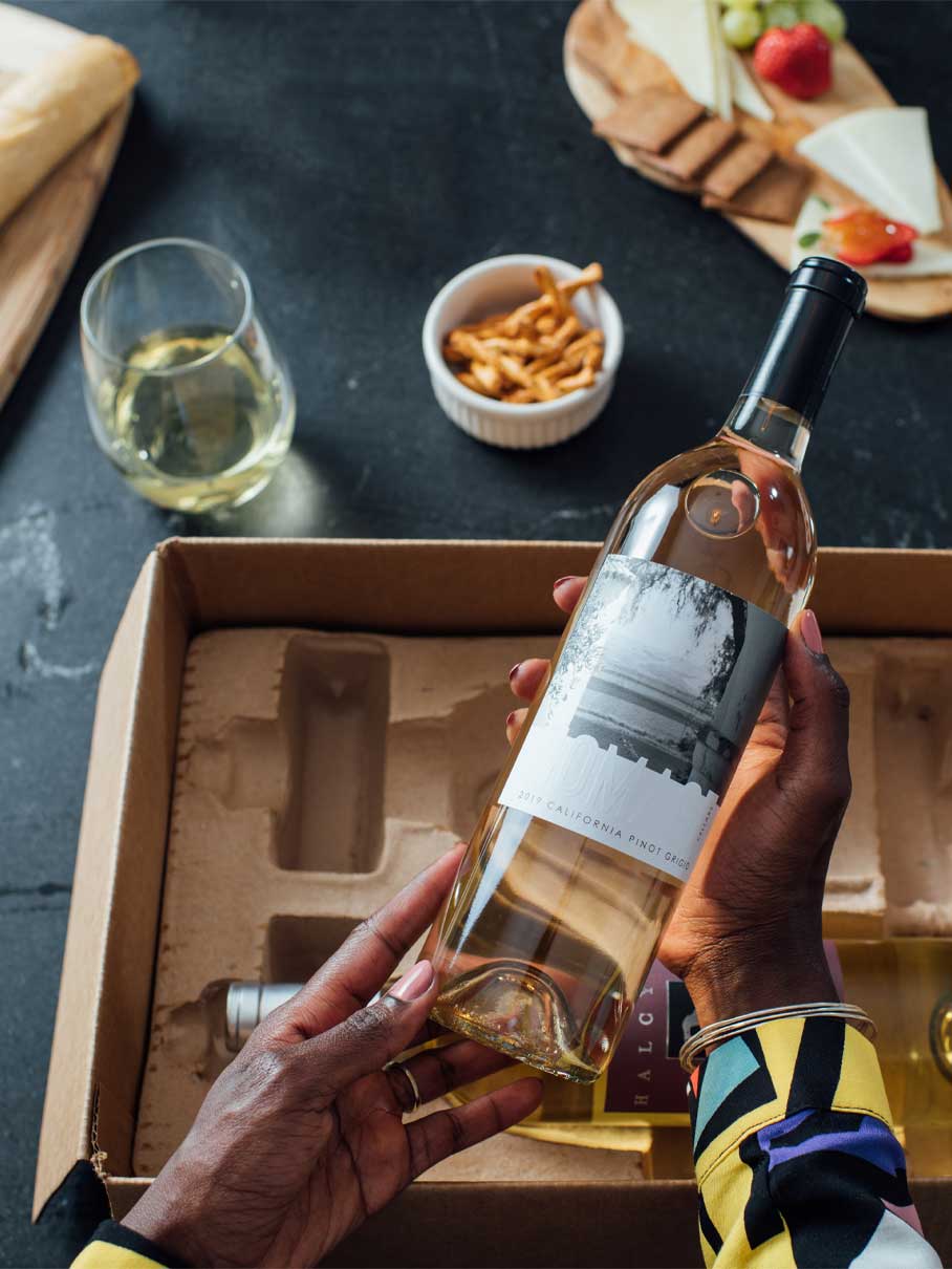 Woman's hands holding a bottle of white wine over a box laid out on a kitchen counter.