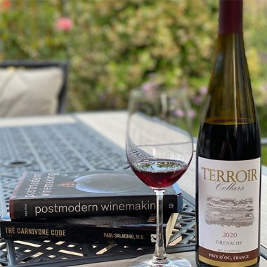 A bottle of Terroir Cellars 2020 Grenache next to a filled glass next to some books on an outdoor patio table