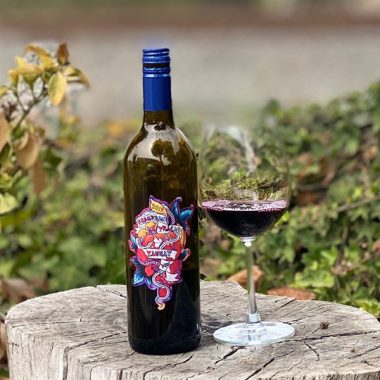 A filled glass of Suburban Fracas 2019 Tannat next to a bottle on a tree stump outdoors