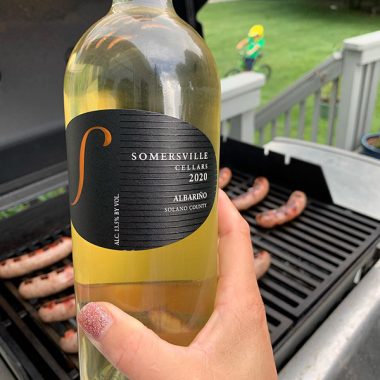A bottle of Somersville Cellars 2020 Albariño being held in front of a grill that has bratwursts grilling on it