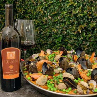 A bottle of Milano Cellars 2020 Merlot next to a filled glass paired with paella outdoors