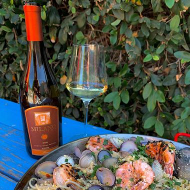 A bottle of Milano Cellars 2020 Trilogy next to a filled glass paired with paella outdoors