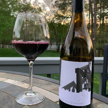 A bottle of Homage Cellars 2019 Petite Sirah and filled glass on a porch table outside