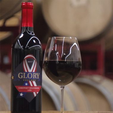Close up of a bottle and filled glass of Glory Cellars 2017 Cabernet Sauvignon in front of some wine barrels