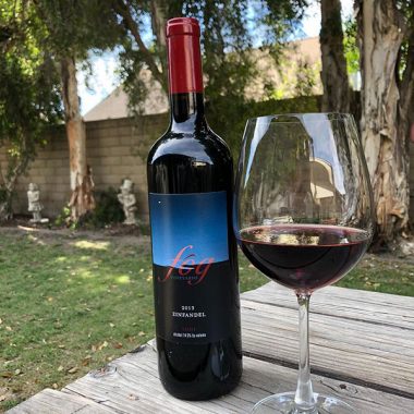 A bottle of Fog Vineyards 2013 Zinfandel next to a filled glass on a picnic table in a backyard