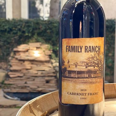 A bottle of Family Ranch 2018 Cabernet Franc on a wine barrel outdoors in front of a beautiful fountain