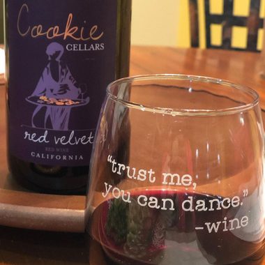A close up of a wine glass that says "trust me you can dance -wine" next to a bottle of Cookie Cellars Red Velet on a kitchen table