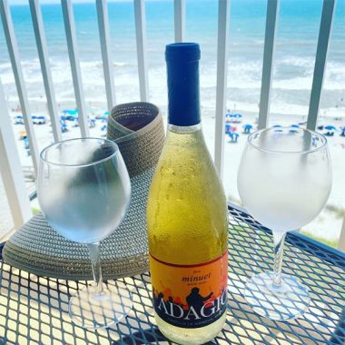 A chilled bottle of Adagio Minuet next to chilled wine glasses and a beach hat on a hotel balcony overlooking the ocean