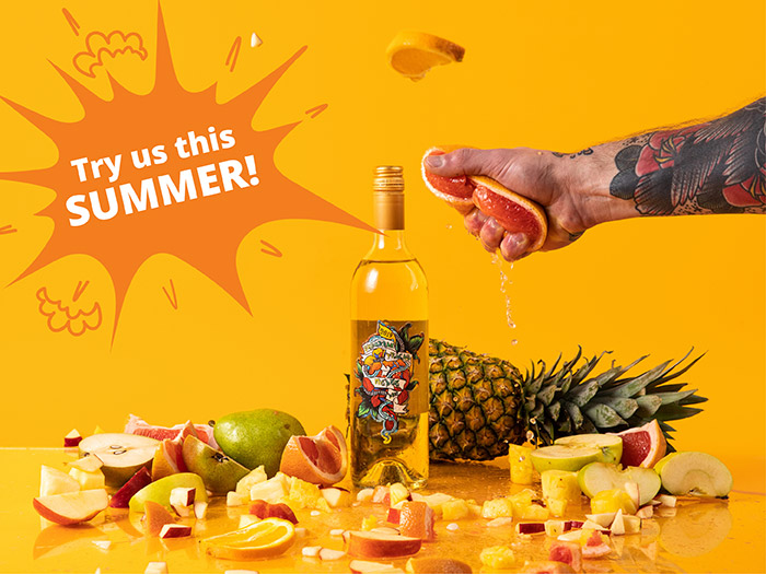 Outstretched arm squeezing grapefruit, picture with Suburban Fracas Melee wines and tropical fruits with the text overlay: try us this summer!